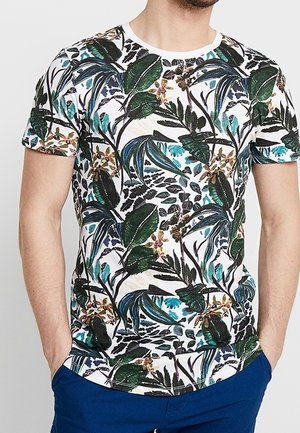 Homme portant un t-shirt blanc à manches courtes avec un imprimé dense de feuilles tropicales vertes et bleues ainsi que des fleurs, associé à un pantalon bleu foncé.