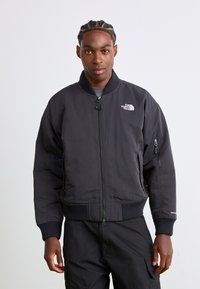 Veste bombardier noire avec fermeture éclair à l'avant, poignets et ourlet côtelés, avec un logo sur la poitrine et une poche latérale. Texture lisse, design isolé.