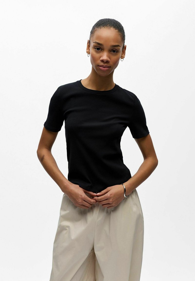 Jeune femme aux cheveux noirs tirés en arrière, portant un haut noir à manches courtes et un pantalon beige, se tenant devant un fond blanc uni.