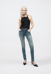 Μαύρο εφαρμοστό τοπ χωρίς μανίκια, μπλε τζιν παντελόνι στενό με διαβάθμιση χρώματος, μυτερές μαύρες γόβες και κολιέ choker με κόκκινη λεπτομέρεια, ποζάροντας με τα χέρια στις τσέπες.