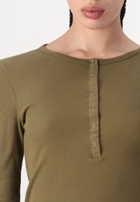 Oliven grønn ribbet langermet topp med rund hals og en rad av fem metallknapper i midten foran. Jevn tekstur.