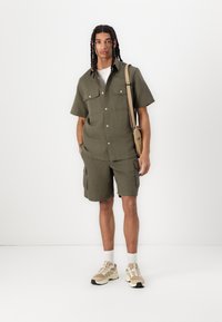 Khaki kortærmet skjorte og cargo shorts sæt i et tekstureret stof, suppleret med en lys beige messenger bag og sneakers.