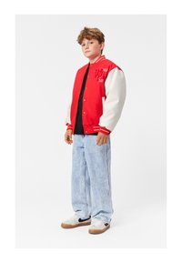 Crvena i bijela vafl jakna s kopčama, s izvezenim detaljima i prugama. Nosena preko crne majice s laganim plavim širokim trapericama i tenisicama.