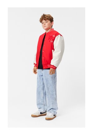 Giacca varsity rossa e bianca con bottoni a pressione, con ricami e strisce. Indossata sopra una maglietta nera con jeans larghi blu chiaro e sneaker.
