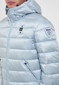 Giacca a vento azzurro chiaro con cappuccio, design trapuntato, tasche laterali con zip e patch con logo sulla spalla sinistra. Tessuto liscio, materiale leggero.