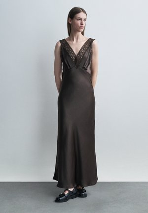 Femme debout portant une longue robe en satin marron foncé avec un col en V en dentelle et des mocassins en cuir noir sur un sol gris contre un mur gris clair.