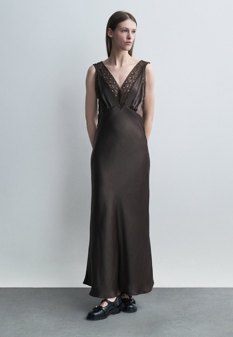 Γυναίκα που στέκεται φορώντας ένα μακρύ σκοτεινό καφέ σατέν φόρεμα με V-λαιμόκοψη από δαντέλα και μαύρα δερμάτινα λοφερς πάνω σε γκρι πάτωμα μπροστά από ανοιχτό γκρι τοίχο.