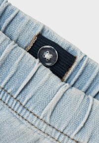Ceinture en denim bleu clair avec une finition texturée, des bords effrangés et un bouton en plastique transparent au centre avec un élastique noir en dessous.