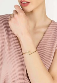 Brazalete de oro con diseño en forma de U y pequeño acento de cristal, llevado en una mano. El fondo presenta una tela plisada suave de color rosa claro.