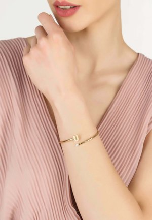 Bracciale d'oro con design a forma di U e piccolo cristallo decorativo, indossato su una mano. Lo sfondo presenta un tessuto morbido e plissettato di colore rosa chiaro.
