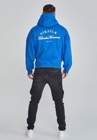Felpa con cappuccio blu con testo bianco sulla schiena, abbinata a jeans cargo neri e sneakers bianche, vista da dietro.