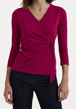 Kobieta ubrana w różowy, długi rękaw, kopertowy top z ozdobą w postaci wiązania z boku, zestawiony z ciemnoniebieskimi dżinsami na jednolitym tle.