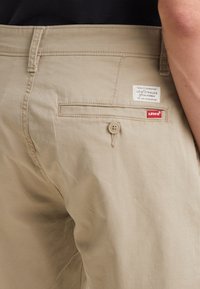 Pantaloni beige con una vestibilità rilassata, dotati di chiusura con bottone sulla tasca posteriore e etichette con marchio. Il tessuto appare leggero e resistente alle pieghe.