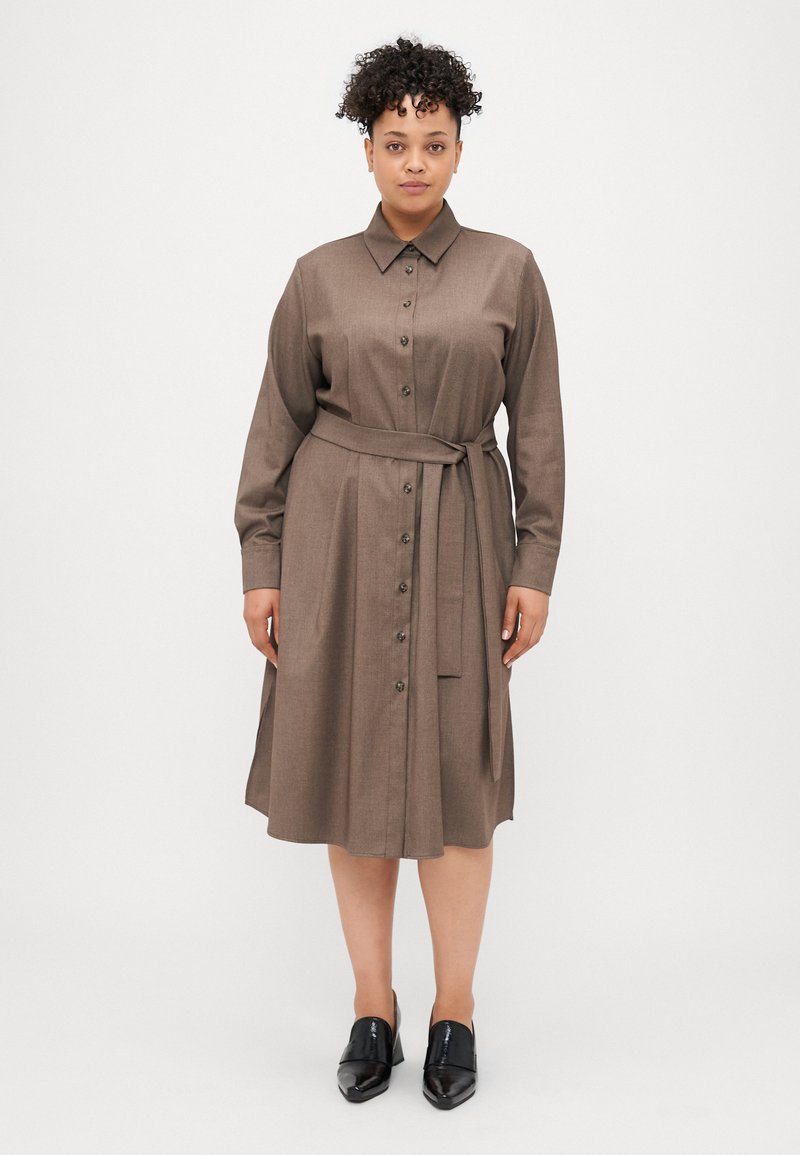 Barna gombos ruha hosszú ujjal, övvel a deréknál, térd alatt végződik, sima textúrával és enyhe fényességgel. Fekete cipővel viselve.