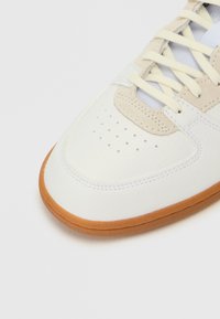 Hvit lær sneakers med beige semsket detaljer, rund tå, perforerte detaljer på tåboksen, og en gummigummisåle.