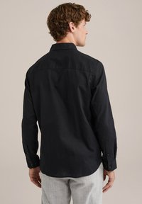 Črna dolga majica z dolgimi rokavi in ovratnikom ter gumbnimi manšetami, ki se ponaša z gladko teksturo in ravnim robom, oblečena z svetlo sivo karirasto hlačami.