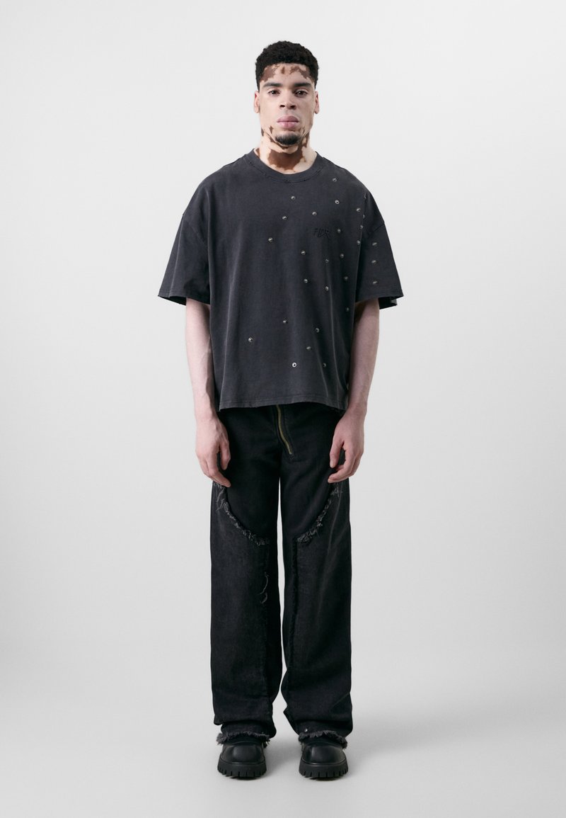 Üks suur must särk, millel on kulunud tekstuur ja metallaktsendid, on seotud vaba mustade teksadega, millel on rebenditega servad ja kollane tõmblukk. Mustad kingad.