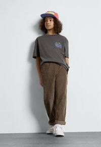 Szary graficzny t-shirt z logo, brązowe spodnie z korduroy i białe trampki. Czapka w kolorach niebieskim, czerwonym i żółtym.