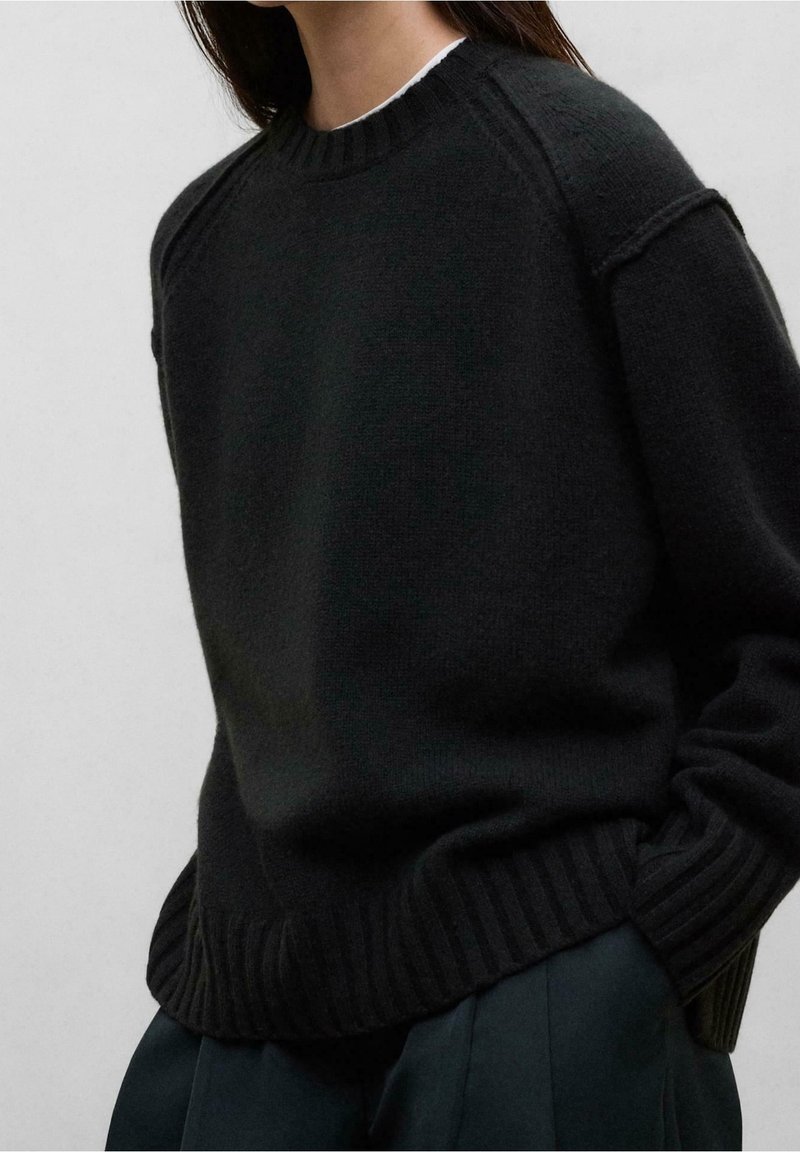 Pull noir en maille avec un col et un ourlet côtelés. Présente une coupe décontractée et des coutures d'épaule tombantes. Texture douce, design minimaliste.
