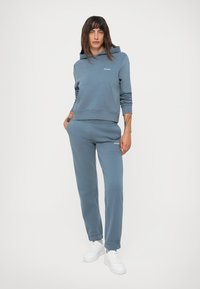 Felpa con cappuccio in cotone blu con tasca frontale, abbinata a pantaloni della tuta coordinati. Presenta polsini a costine e sneakers bianche. Design semplice.