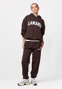 Tânără în picioare, purtând hanorac maro închis și pantaloni de trening cu textul „LAMANIA”, adidași albi și cercei rotunzi argintii.