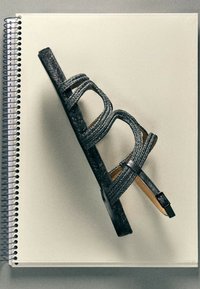 Sandale plate noire tressée avec bride de cheville et boucle reposant sur un carnet à spirale fermé avec une couverture blanche unie.
