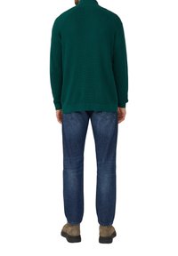 Dunkelgrüner Strickpullover mit hohem Kragen, kombiniert mit blauen Jeans und braunen Schuhen, von hinten betrachtet.