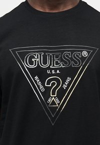 Μαύρο μπλουζάκι με ασημένιο τριγωνικό λογότυπο Guess, με μεγάλο ερωτηματικό και τα κείμενα "U.S.A", "Washed" και "Jeans".