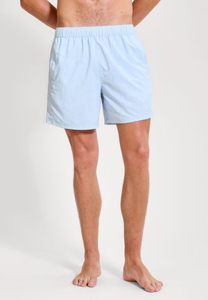 Homme portant un short bleu clair à taille élastique, debout pieds nus devant un fond clair uni.