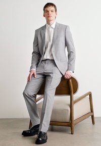 Completo su misura grigio chiaro con giacca a colletto a scendere, pantaloni slim-fit e camicia bianca, abbinato a scarpe in pelle nere.