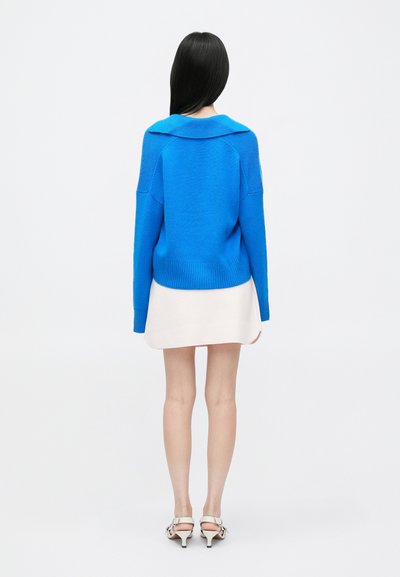 Niebieski sweter dzianinowy z szerokim kołnierzem, luźny krój, prążkowany dół i długie rękawy, zestawiony z jasną spódnicą i butami na obcasie.