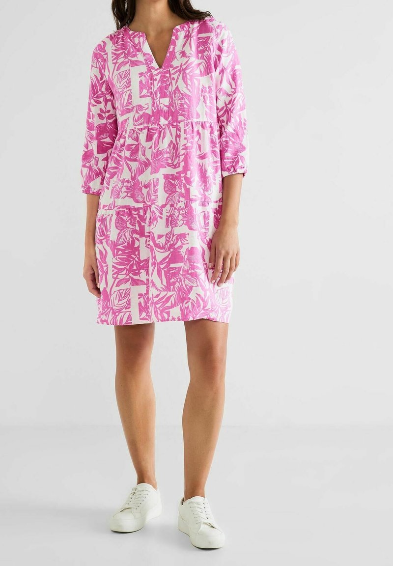 Femme portant une robe fleurie rose et blanche à longueur genou avec des manches trois-quarts et des baskets blanches, debout devant un fond uni.