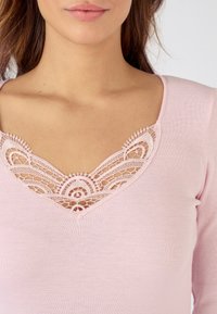 Maglione a maniche lunghe di colore rosa con dettaglio in pizzo a V, caratterizzato da motivi intricati, circondato da un tessuto di cotone morbido.