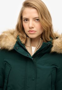 Parka verde con capucha de pelo, cierre de cremallera y bolsillos laterales. Llevada sobre un suéter beige, con un acabado liso y texturizado.