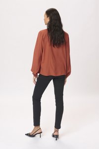 Roestkleurige blouse met lange mouwen en gestructureerde stippen, gecombineerd met zwarte skinny jeans en zwarte slingback hakken, weergegeven vanuit de achterkant.