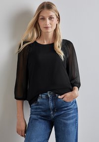 Blusa nera con maniche trasparenti a tre quarti, tessuto testurizzato, scollatura rotonda, abbinata a jeans in denim blu, tasca frontale visibile.