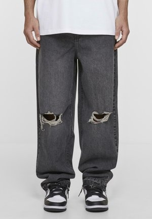 Baggy mørkegrå denimjeans med slidte knæ, frynset kant og en glat tekstur, parret med sort og hvide sneakers.