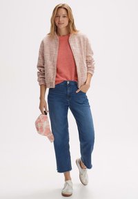 Lichtroze bomberjack van textuurstof, koraalkleurig T-shirt, blauwe cropped jeans en witte sneakers; houdt een roze tie-dye pet vast.