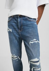 Niebieskie dżinsy jeansowe o wąskim kroju, z licznymi przetarciami, delikatnym postrzępieniem oraz miedzianym przeszyciem wzdłuż szwów.