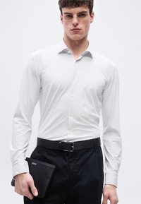 Νεαρός άνδρας που φοράει λευκό πουκάμισο με μοτίβο και μαύρο παντελόνι, κρατώντας μια μαύρη τσάντα χειρός με το λογότυπο Hugo στο αριστερό χέρι.