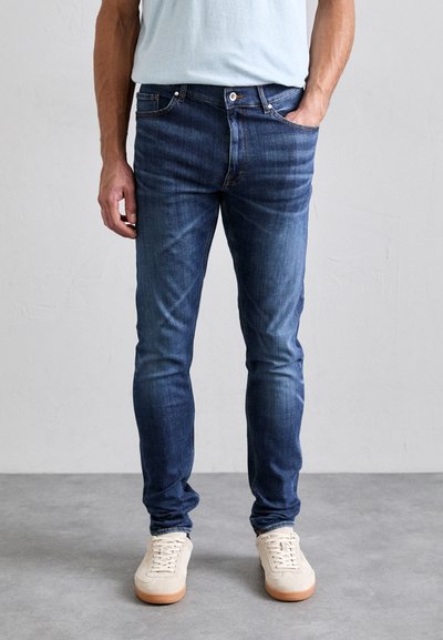 Sötétkék, slim fit farmernadrág, öt zsebes kialakítással, enyhe fakulással és egyetlen gombbal záródik, krémszínű sportcipővel párosítva.