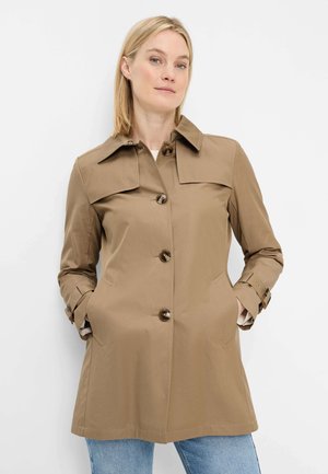 Woman wearing a light brown, buttoned trench coat with hands in pockets, standing against a plain white background.