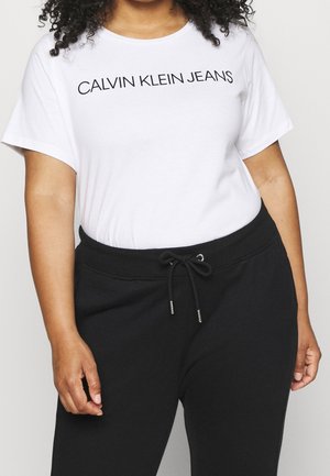 Nainen, jolla on valkoinen Calvin Klein Jeans -T-paita, joka on sullottu mustien vetoketjullisten collegehousujen sisään, seisoo rennosti käsivarret rentoina.