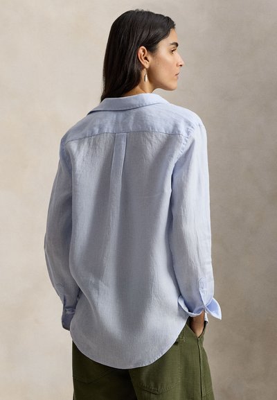 Γυναίκα με σκούρα μαλλιά, που φοράει ανοιχτό μπλε πουκάμισο μακρυμάνικο και χακί παντελόνι, στέκεται με τα χέρια στις τσέπες, γυρισμένη πλάτη στην κάμερα.