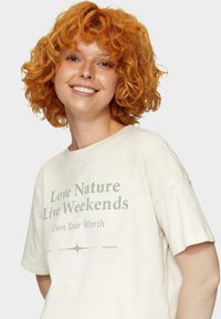 Lichtbeige t-shirt van katoen met groene tekst: "Hou van de Natuur Leef in het Weekend" en "Neem je Waardering op", met korte mouwen en een relaxte pasvorm.