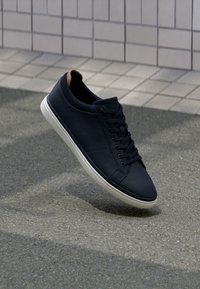 Marineblaue Leder-Sneaker mit glatter Textur, weißer Gummisohle und traditionellem Schnürdesign. Mit einem braunen Akzent an der Ferse.