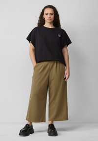 T-shirt en coton noir à manches courtes et avec un petit graphique. Pantalons larges vert olive en tissu lisse. Chaussures noires épaisses.