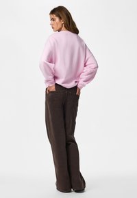 Femme debout de dos, portant un haut à manches longues rose clair et un pantalon ample marron foncé sur un fond blanc uni.