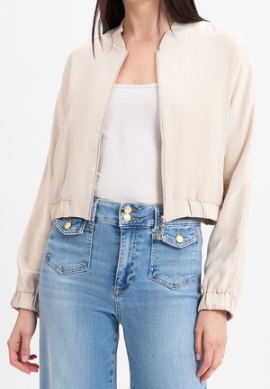 Femme portant une veste courte beige zippée sur un débardeur blanc, associée à un jean taille haute bleu clair avec des boutons dorés.