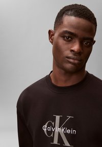 Fiatalember rövid hajjal, fekete Calvin Klein pulóvert viselve, tiszta világosszürke háttér előtt.